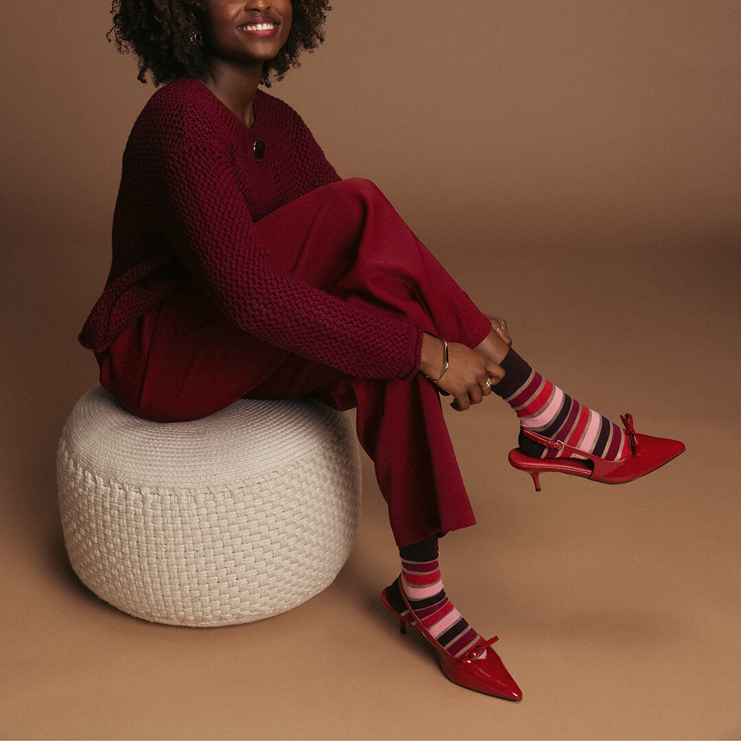 Woman in red outfit with striped socks and red high heels sitting on a white woven stool against a brown background