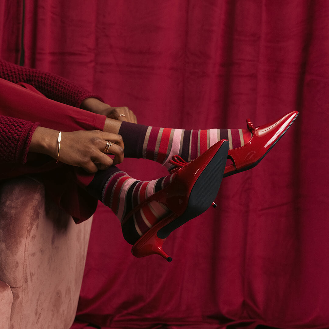 Person wearing red high-heeled shoes and striped socks sitting on a pink couch against a red curtain.