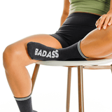 Person wearing black socks with 'BADASS' text, sitting on a chair.