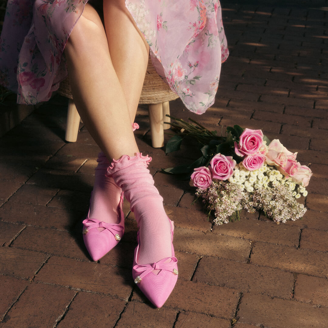 Person wearing pink high heels and pink slouch lettuce-edge socks with bow details on a brick floor, accompanied by a bouquet.