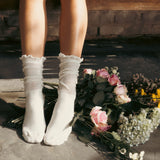 White socks with ruffles worn by a person sitting on a stone surface with flowers.
