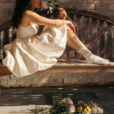 Woman in a white dress and feminine slouch socks sitting on a wooden bench with flowers, surrounded by more flowers on the ground.