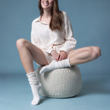 Woman sitting on a woven stool wearing white  sheer slouch socks against a blue background